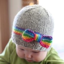 Baby wears simple knitted beanie in neutral color with rainbow stripe above the brim. Affixed to the stripe is a matching knitted rainbow bow.