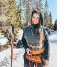 Smiling woman in a snowy wood wears sleeveless sweater has tie at waist, a few buttons at the neck and a cozy hood. Design is Hopi-inspired.