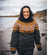 Smiling woman at the beach wears charcoal gray and geometric colorwork sweater. Yoke and a band around each wrist is in geometric colorwork in yellow, red, orange and charcoal.